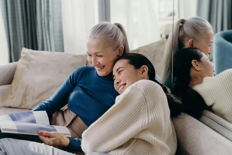 A senior woman with a ponytail wearing a blue sweater and a younger woman in a cream sweater comparing Medicare plans together on a sofa